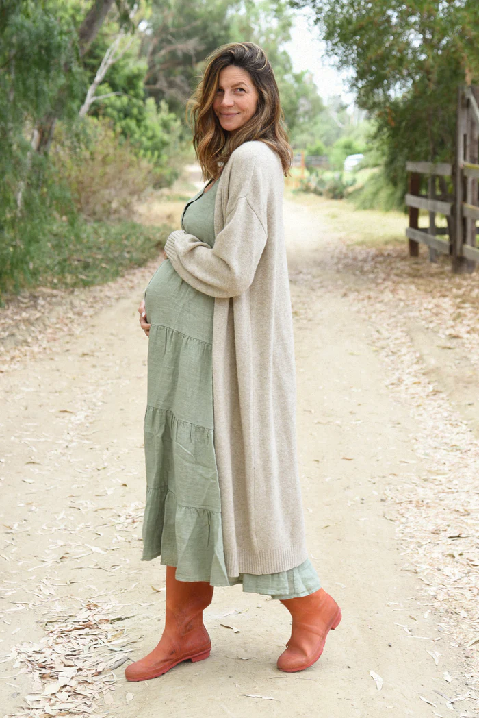 Pregnant woman wearing a sage green Beachwood maternity dress layered with a long beige cardigan, standing on a dirt path with red boots and a serene smile.