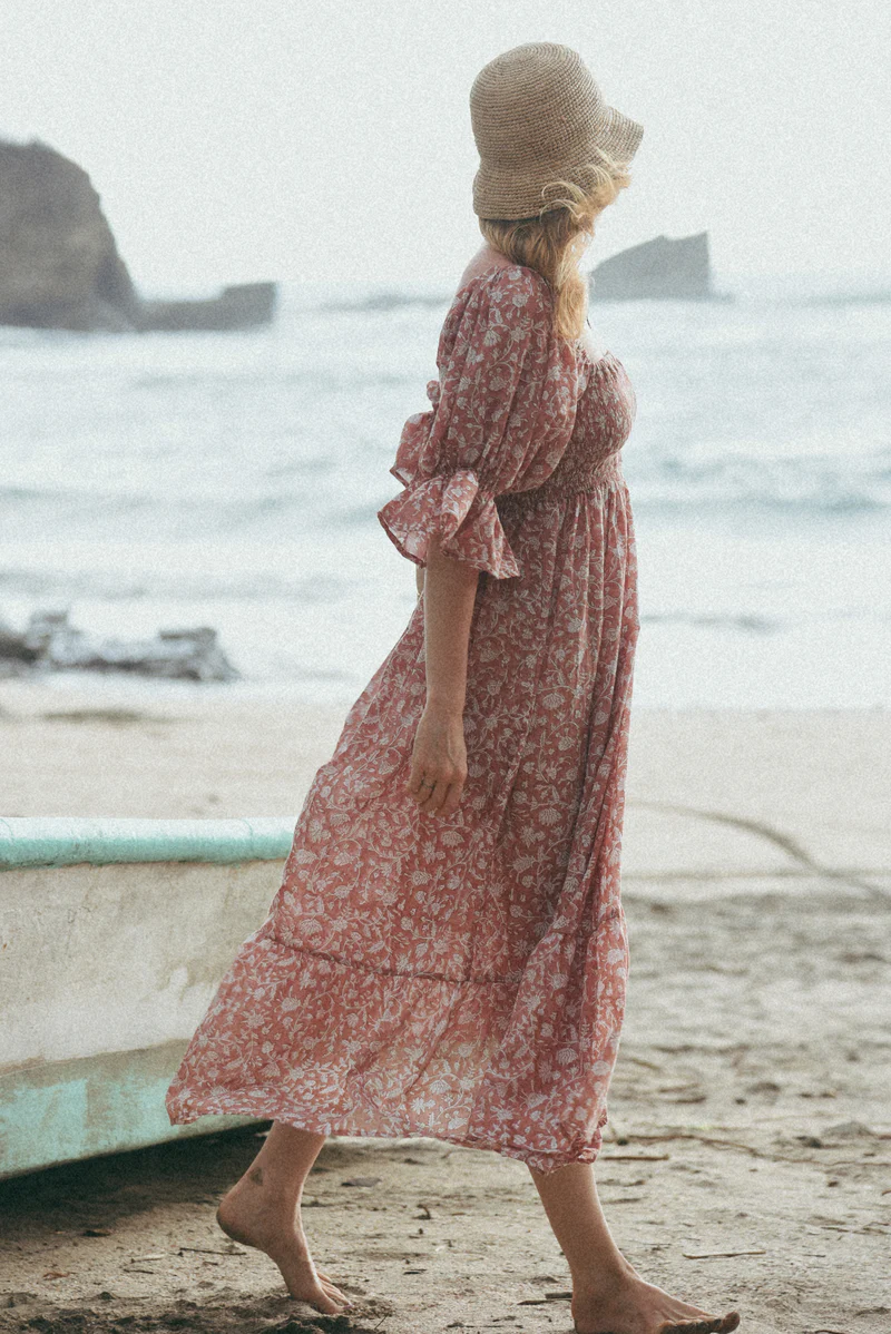 Pregnant woman wearing a flowy, floral Beachwood maternity dress in soft red tones, walking barefoot on a sandy beach with the ocean and rocks in the background.