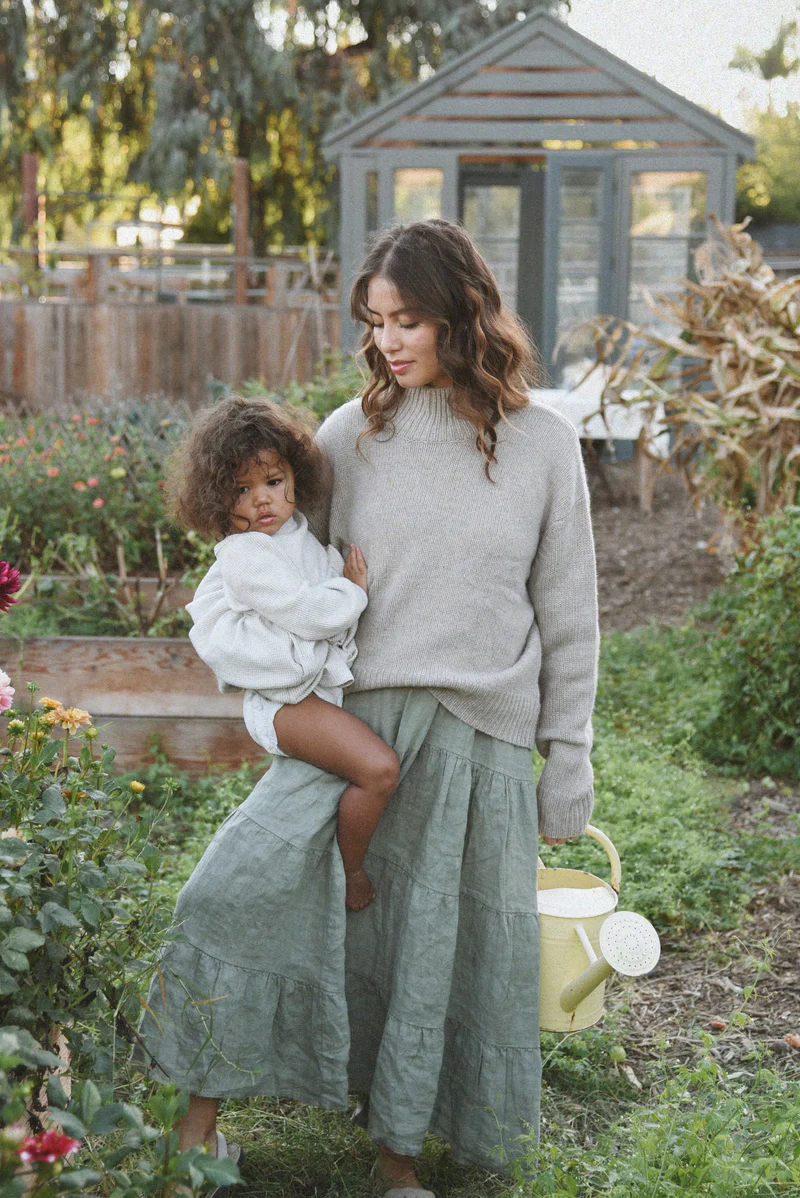 Woman standing in a garden, wearing a light gray Beachwood sweater paired with a flowy sage green skirt, holding a toddler dressed in a cozy white sweater.