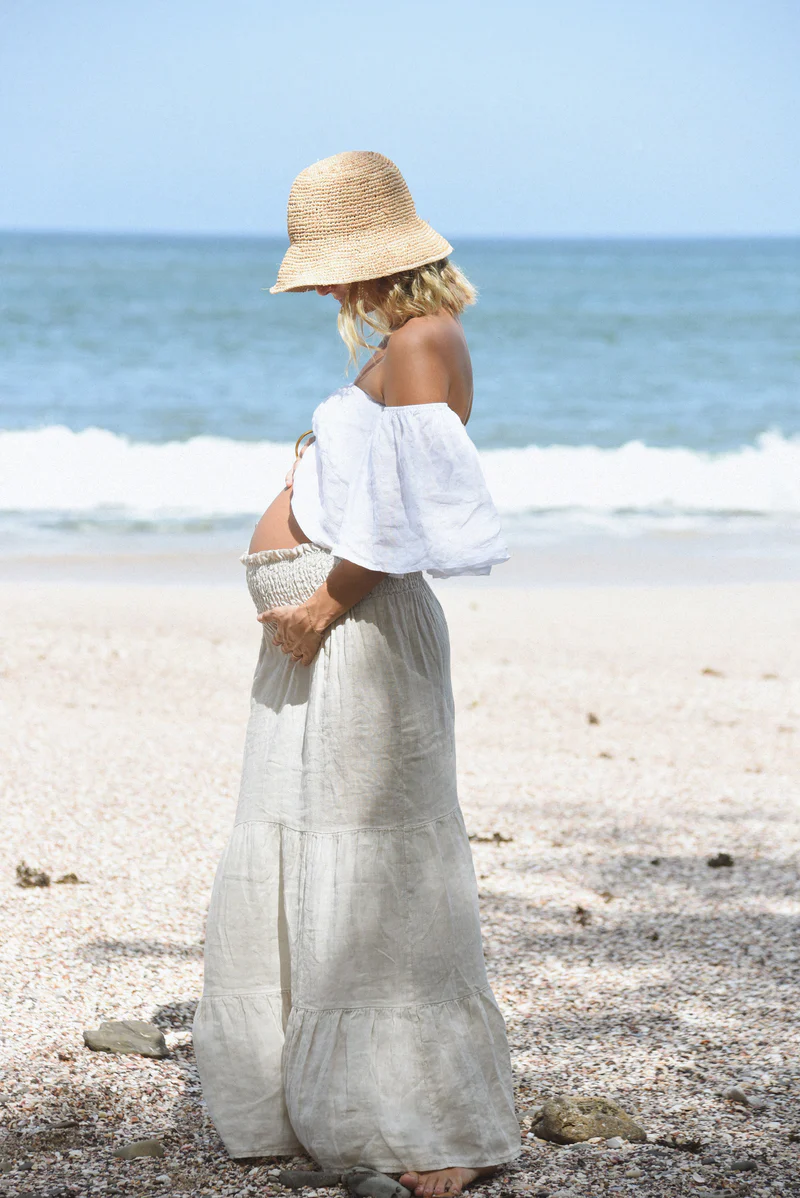 Pregnant woman standing on a sandy beach, wearing a flowy linen Beachwood skirt and an off-shoulder white top, cradling her baby bump under a straw hat.