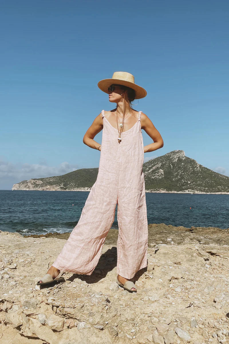 Woman standing confidently on a rocky shore, wearing a flowy pink Beachwood jumpsuit and a straw hat, with a scenic ocean and mountain backdrop under a clear blue sky.