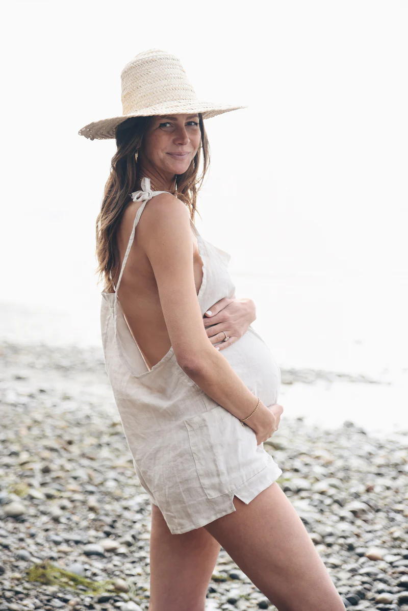 Pregnant woman wearing a light linen Beachwood jumpsuit with tie straps, paired with a wide-brim straw hat, standing on a rocky beach with the ocean breeze in her hair.