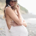 Pregnant woman wearing a light linen Beachwood jumpsuit and a wide-brim straw hat, standing on a rocky beach with the ocean breeze in her hair.