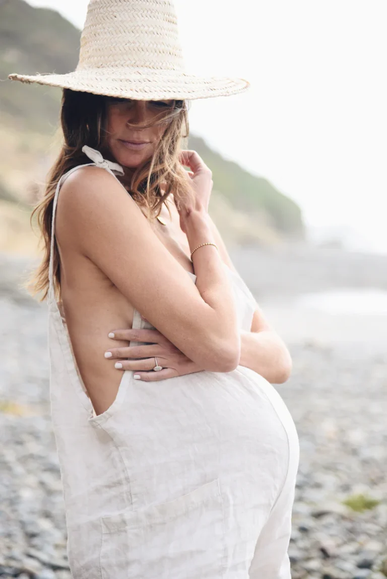 Pregnant woman wearing a light linen Beachwood jumpsuit and a wide-brim straw hat, standing on a rocky beach with the ocean breeze in her hair.