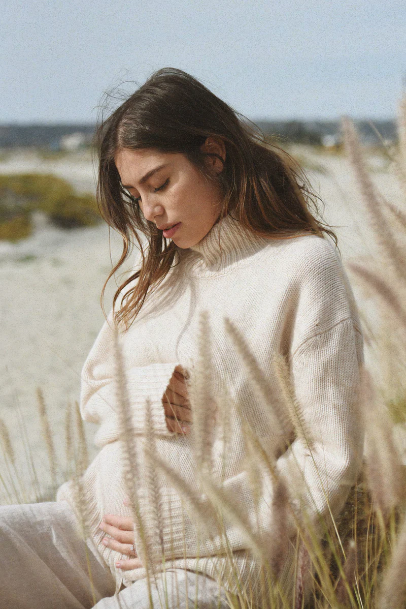 A pregnant woman wearing The Cashmere Carmel Sweater in moon, sitting on a beach surrounded by tall grass.