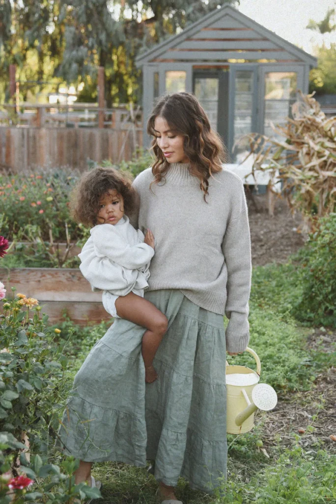 A woman wearing The Cashmere Carmel Sweater in a neutral nutmeg tone, paired with a flowing green skirt, standing in a garden.