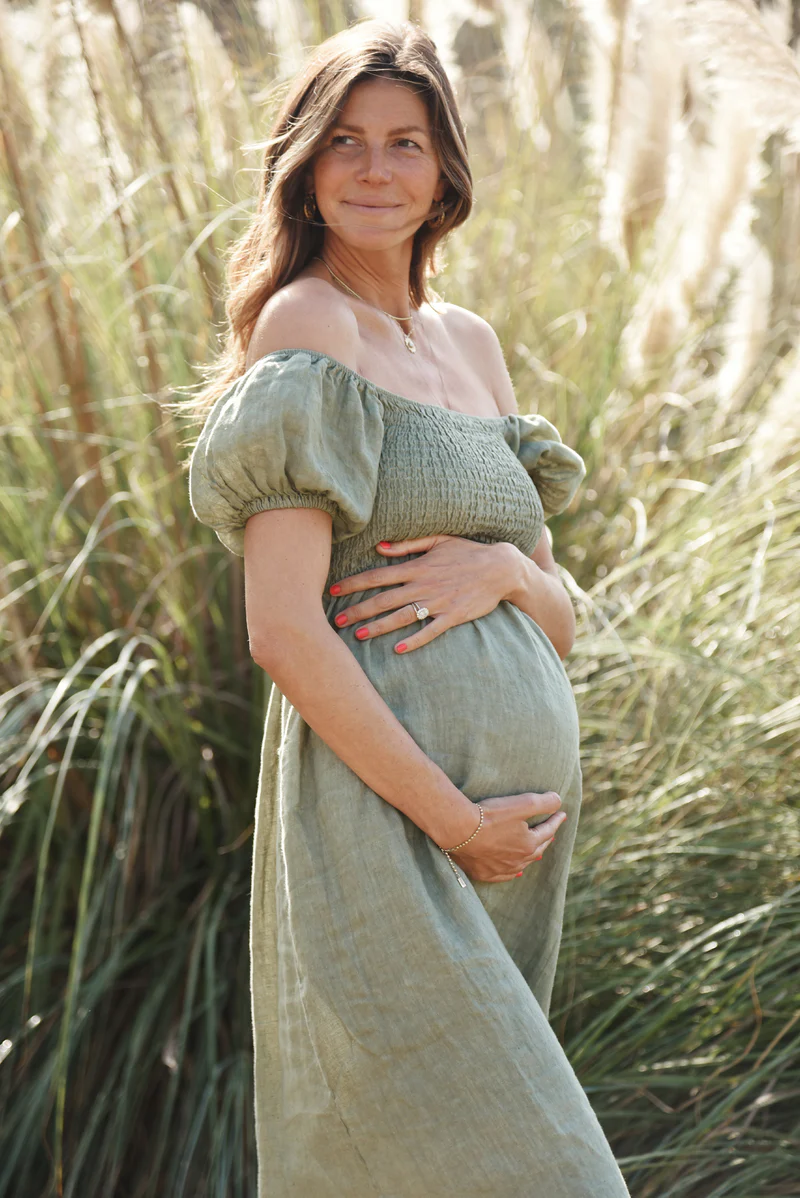 A pregnant woman wearing a sage green Menorca Dress with puffed sleeves, styled off-the-shoulder, standing in tall grass.