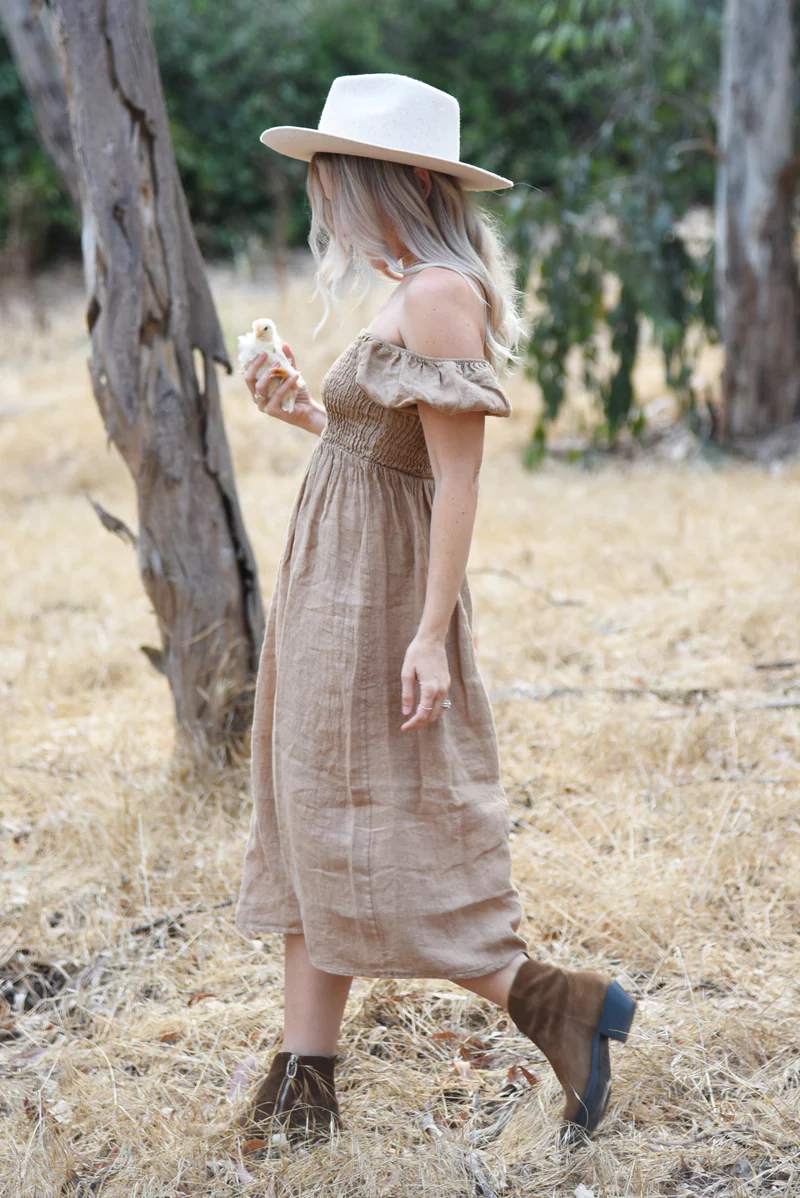 A woman in a rye-coloured Menorca Dress, paired with a hat and boots, walking through a dry autumn field