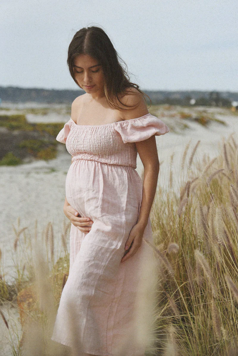 A woman in a blush pink Menorca Dress, designed for pregnancy and postpartum, walking along a beach.